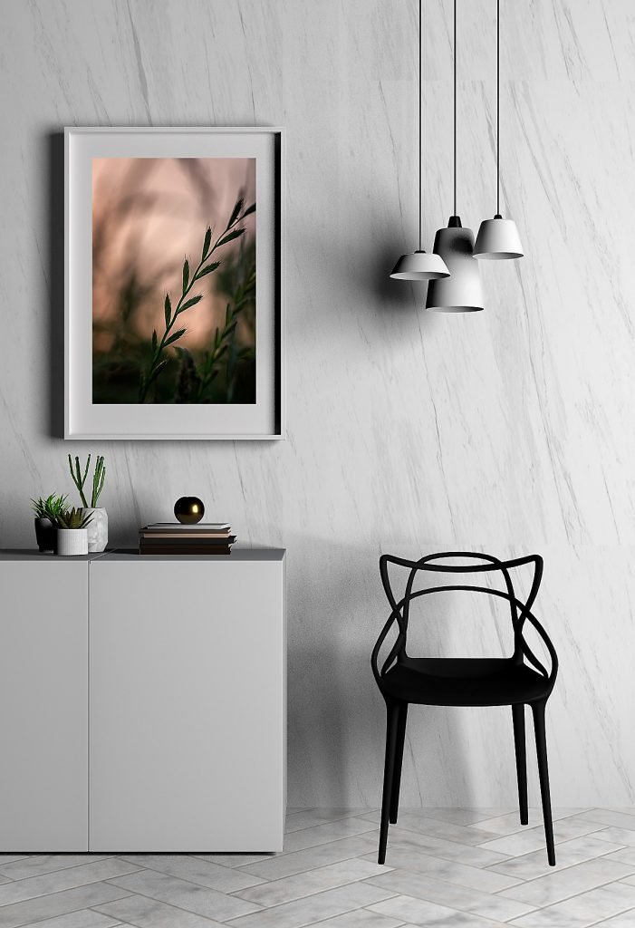 Close-up macro photograph of meadow grass in evening light, framed in a white frame with white mounting, hanging on a wall. A black chair and a white credenza with books and potted succulents are visible nearby.